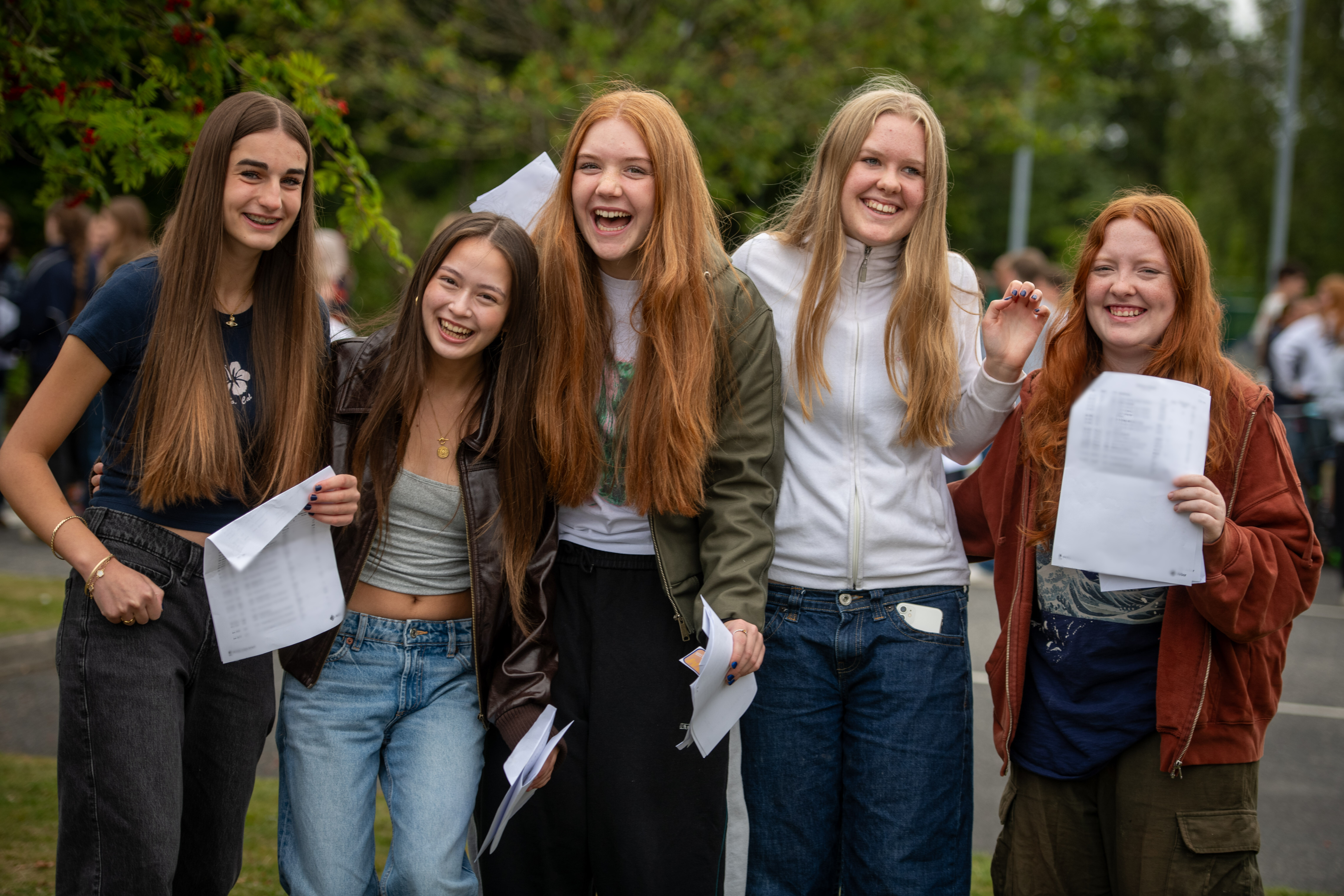 A row of 5 young women smiling as they look at the camera, holding pieces of paper in their hands