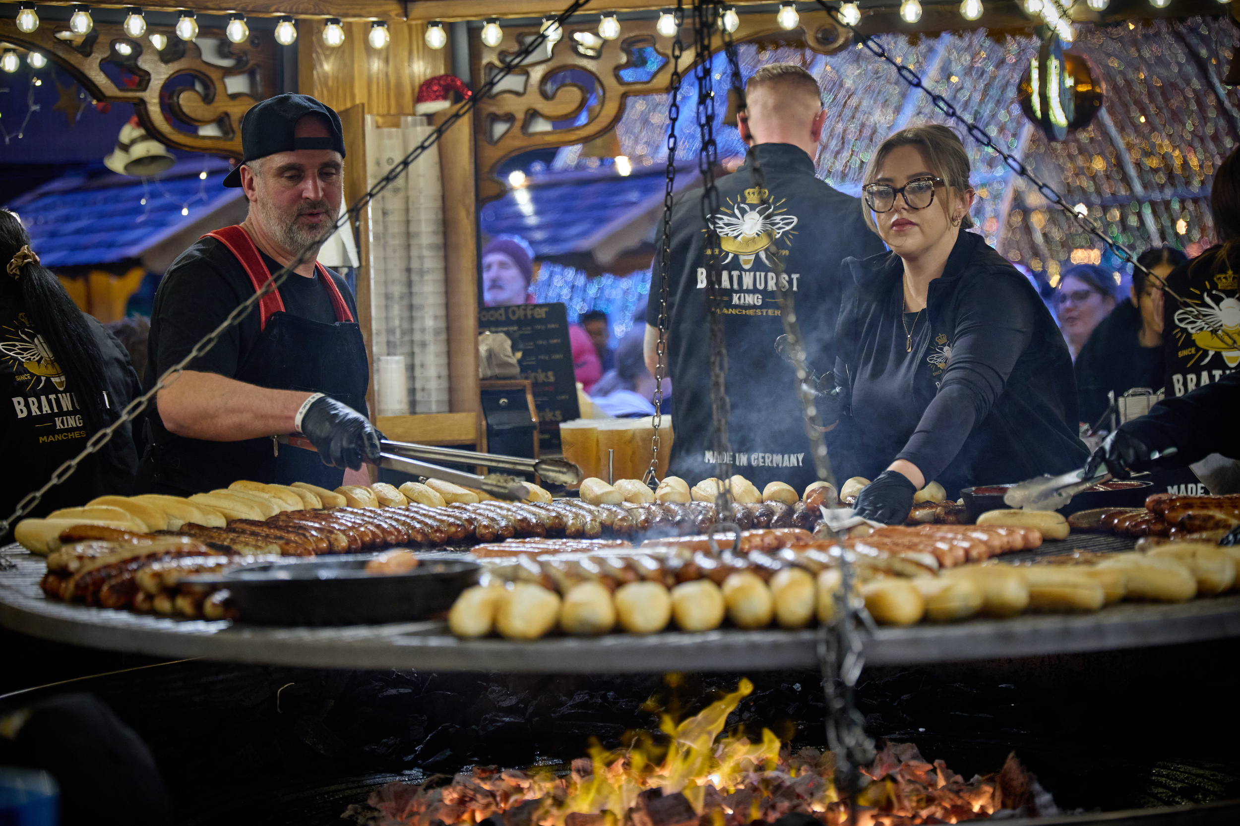 Outside in a market setting. A man and a woman cooking at a big swing grill with sausages and other food on it and a big flame underneath the grill.  Lots of people around.