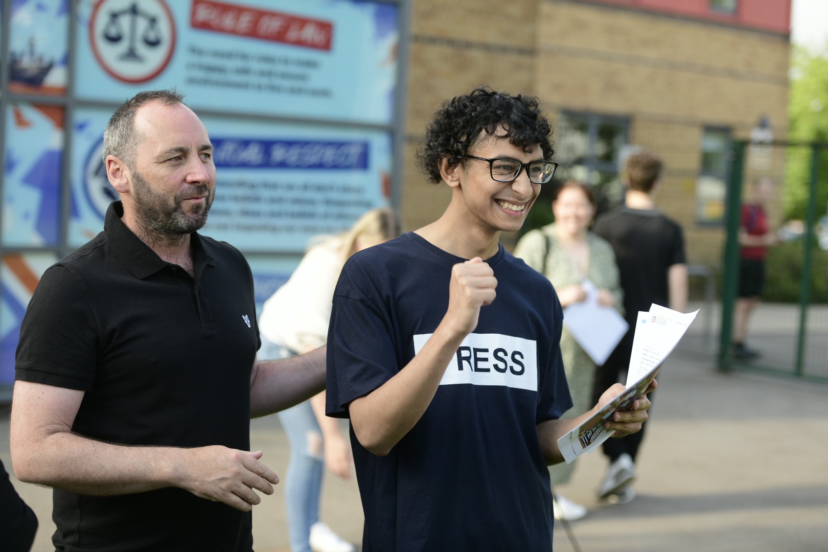 Young person smiling as he holds a piece of paper in one hand and cheers with his other hand as a man stands next to him smiling