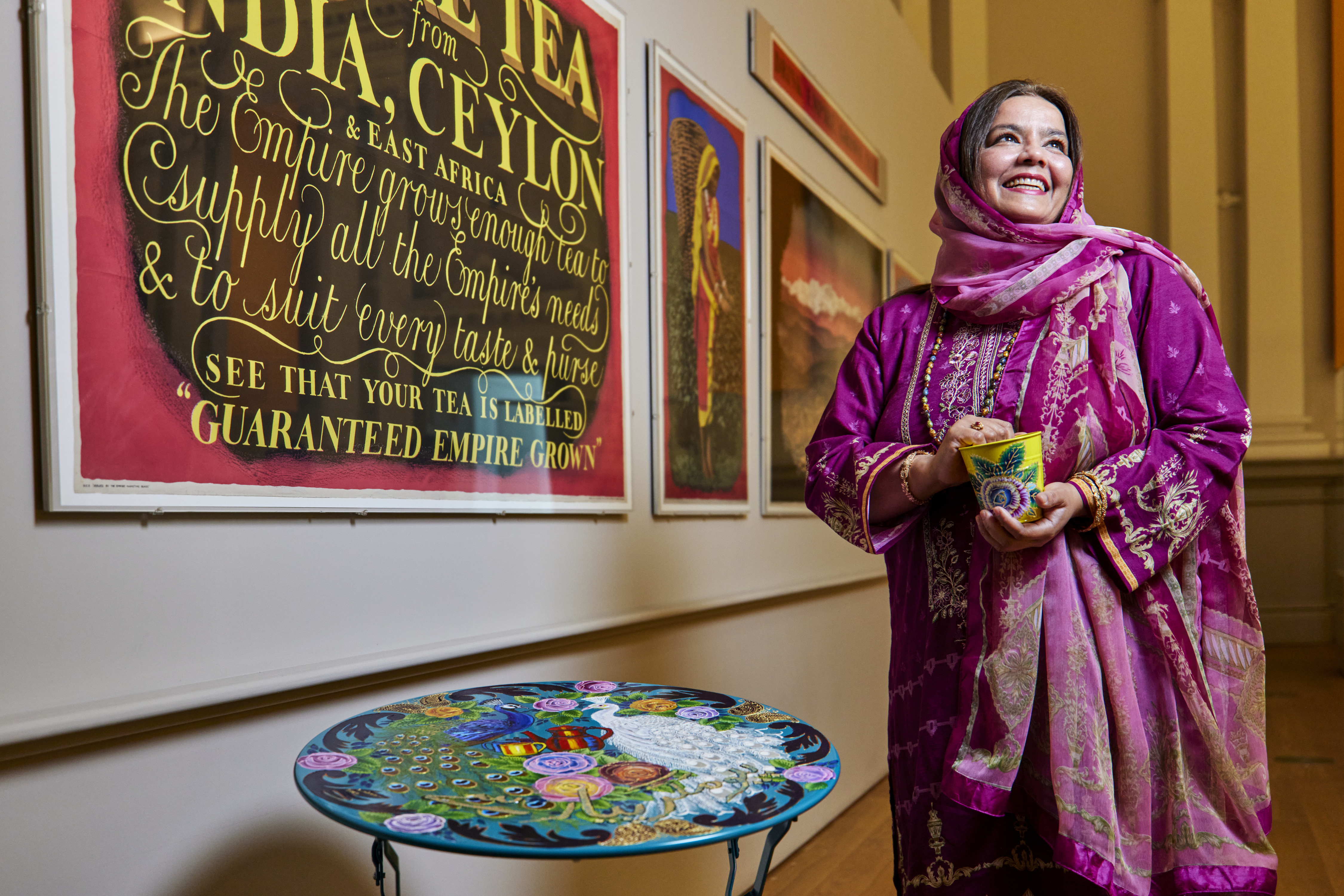 Woman holding a brightly coloured pot in her hands standing next to a brightly painted table, next to a wall with posters on it