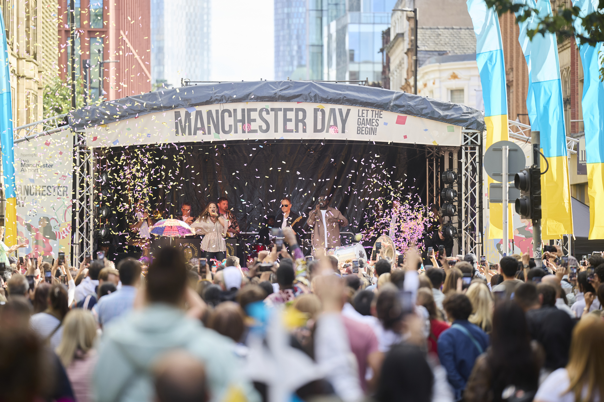 Crowds of people arms in the air watching musicians on a stage with the words Manchester Day above the stage