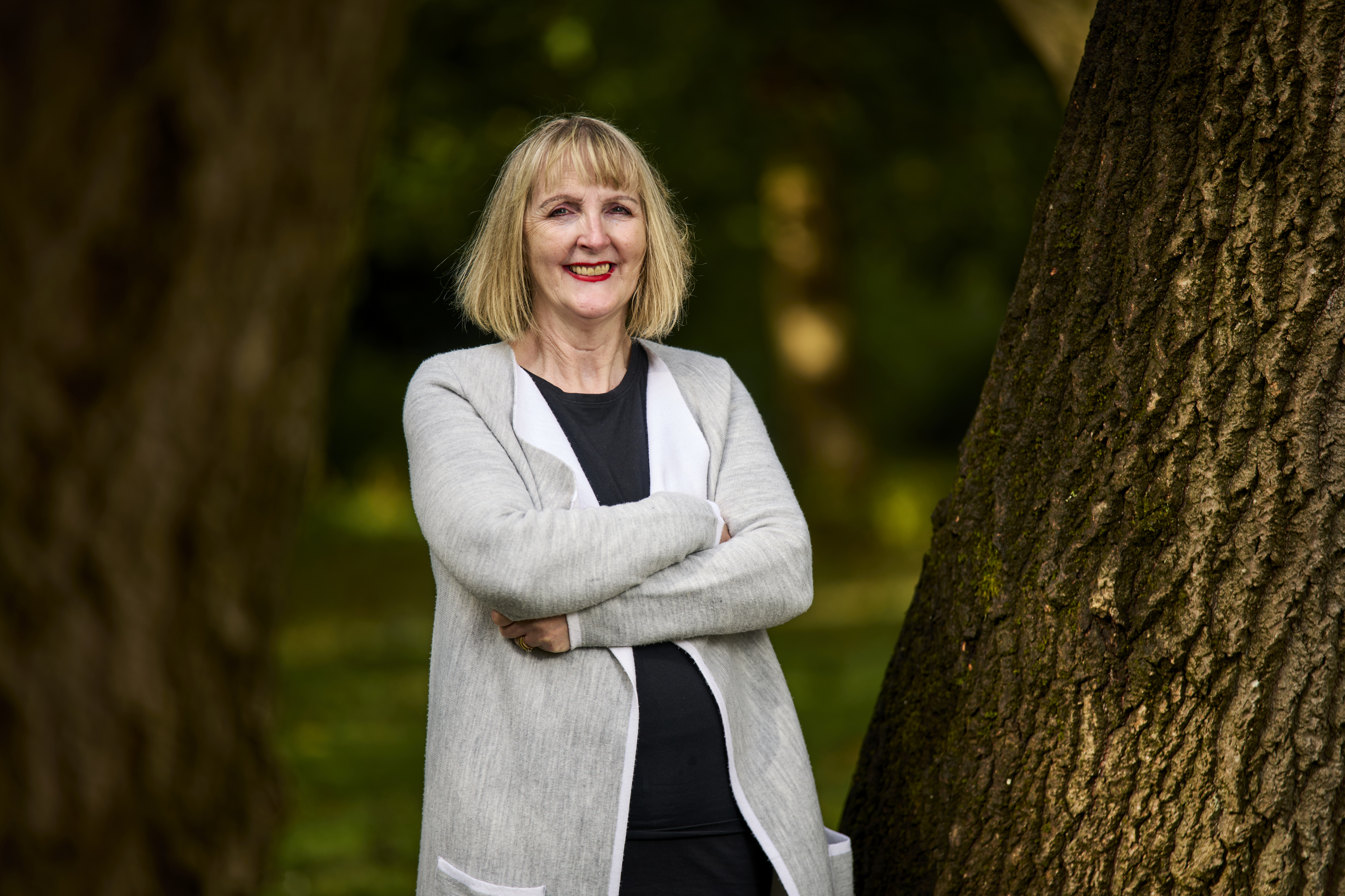 Outside next to trees. 
 A woman stands looking at camera, arms folded, smiling
