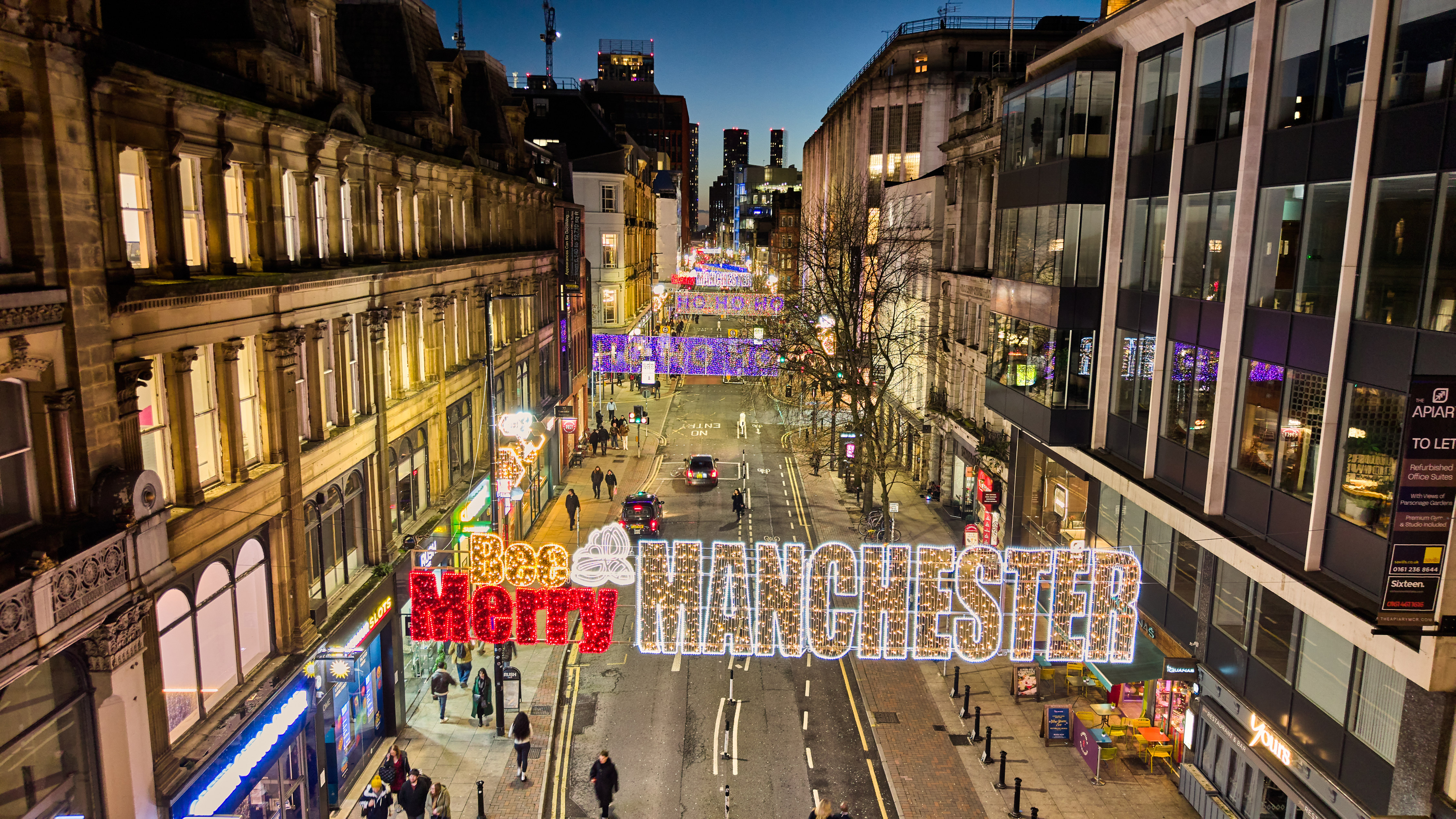 Night time street scene showing Deansgate in Manchester with its wide street and twinkling signs across the street saying Bee Merry Manchester