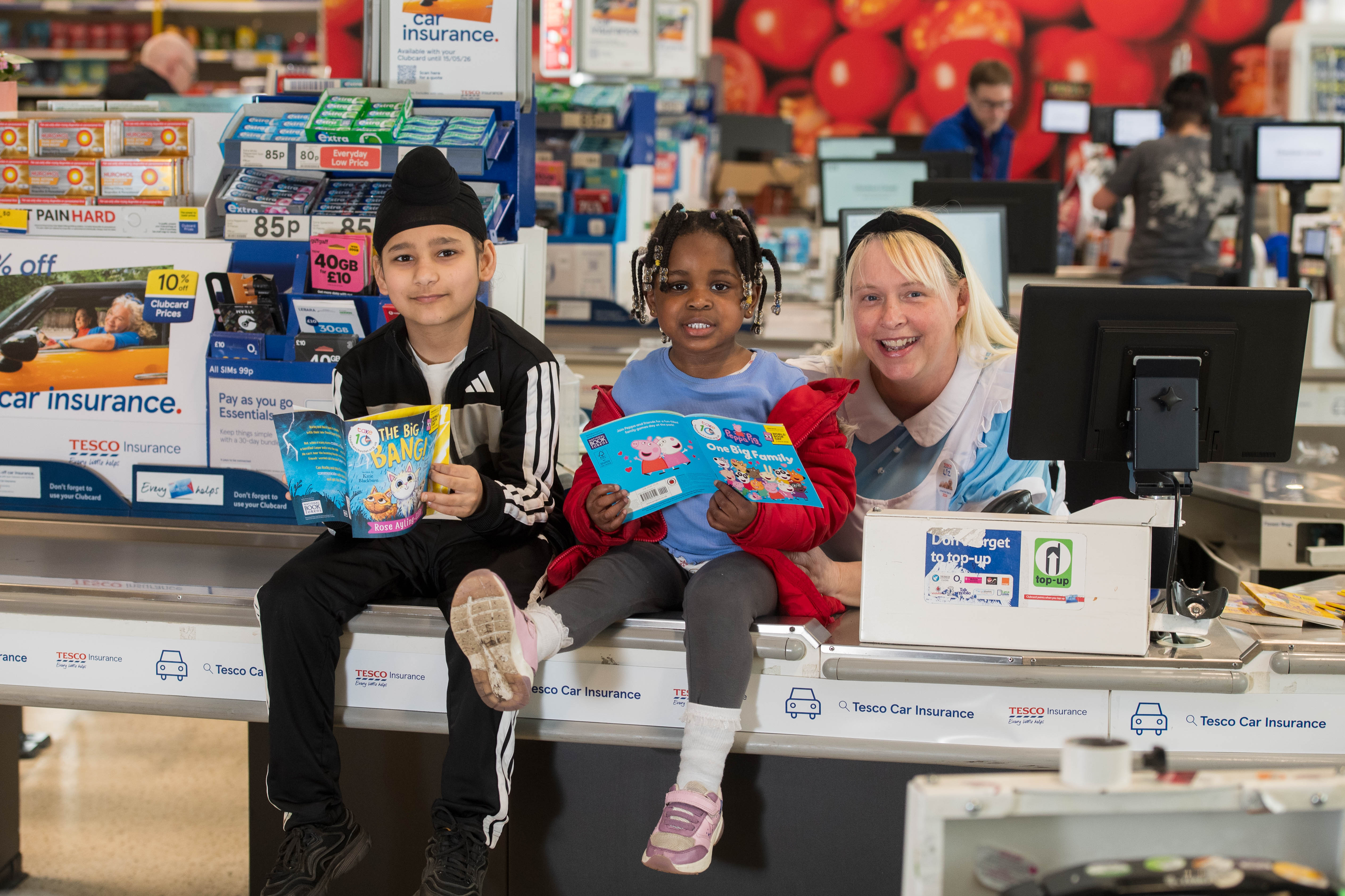 Two children sit on a supermarket checkout conveyer belt holding up a book each and smiling as an adult dressed as Alice in Wonderland leans in next to them