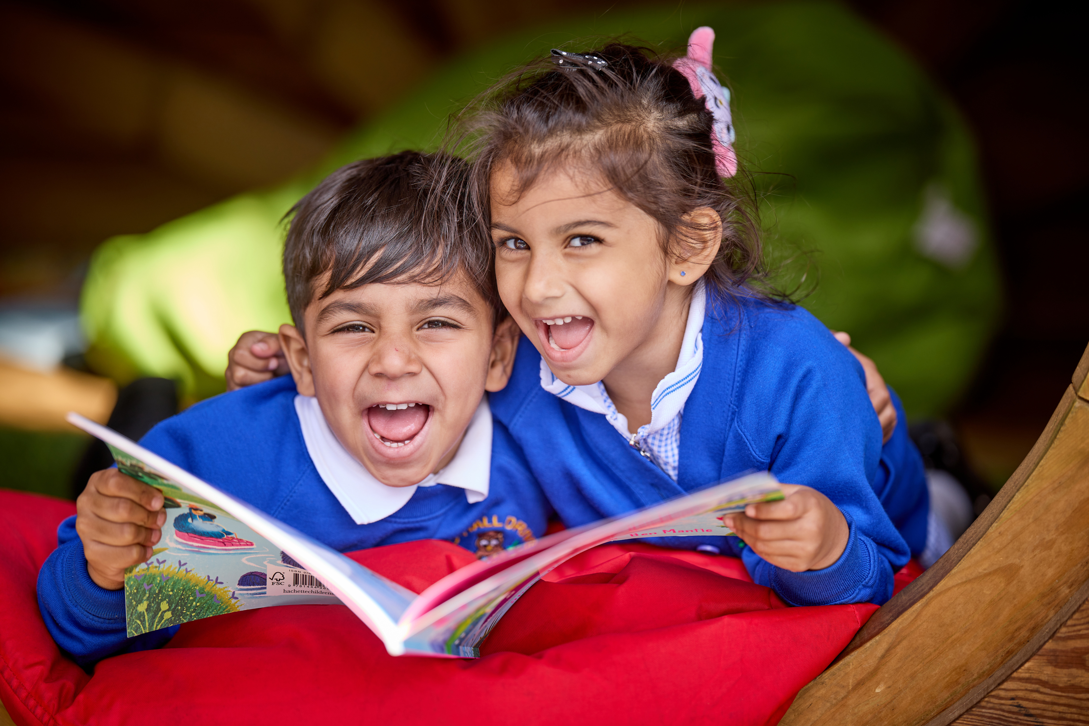 A little boy and girl aged around 4 wearing blue school uniforms, holding a big book between them as they smile at the camera
