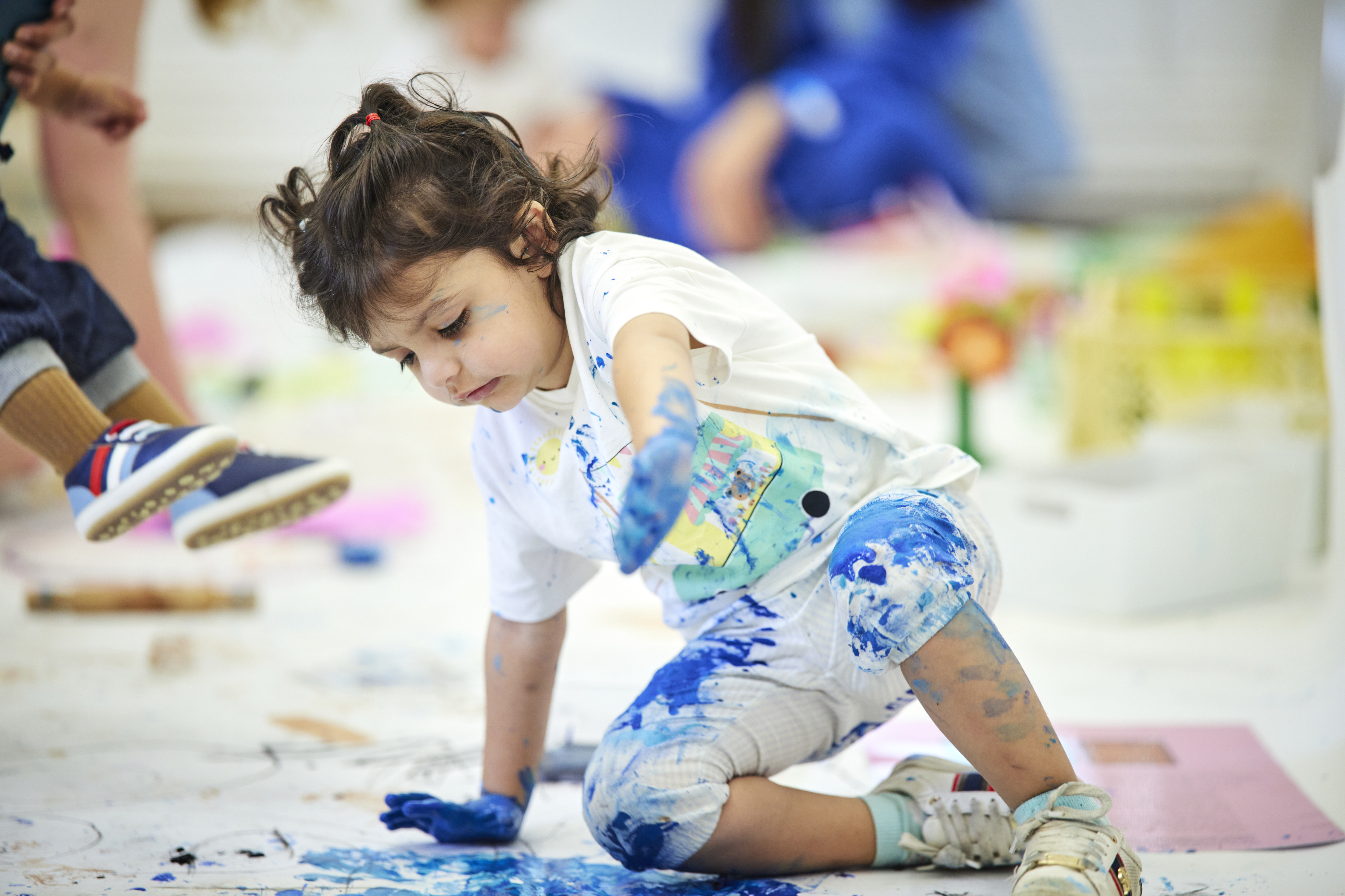 Young child hands and clothes covered in paint splashes, sitting on the floor