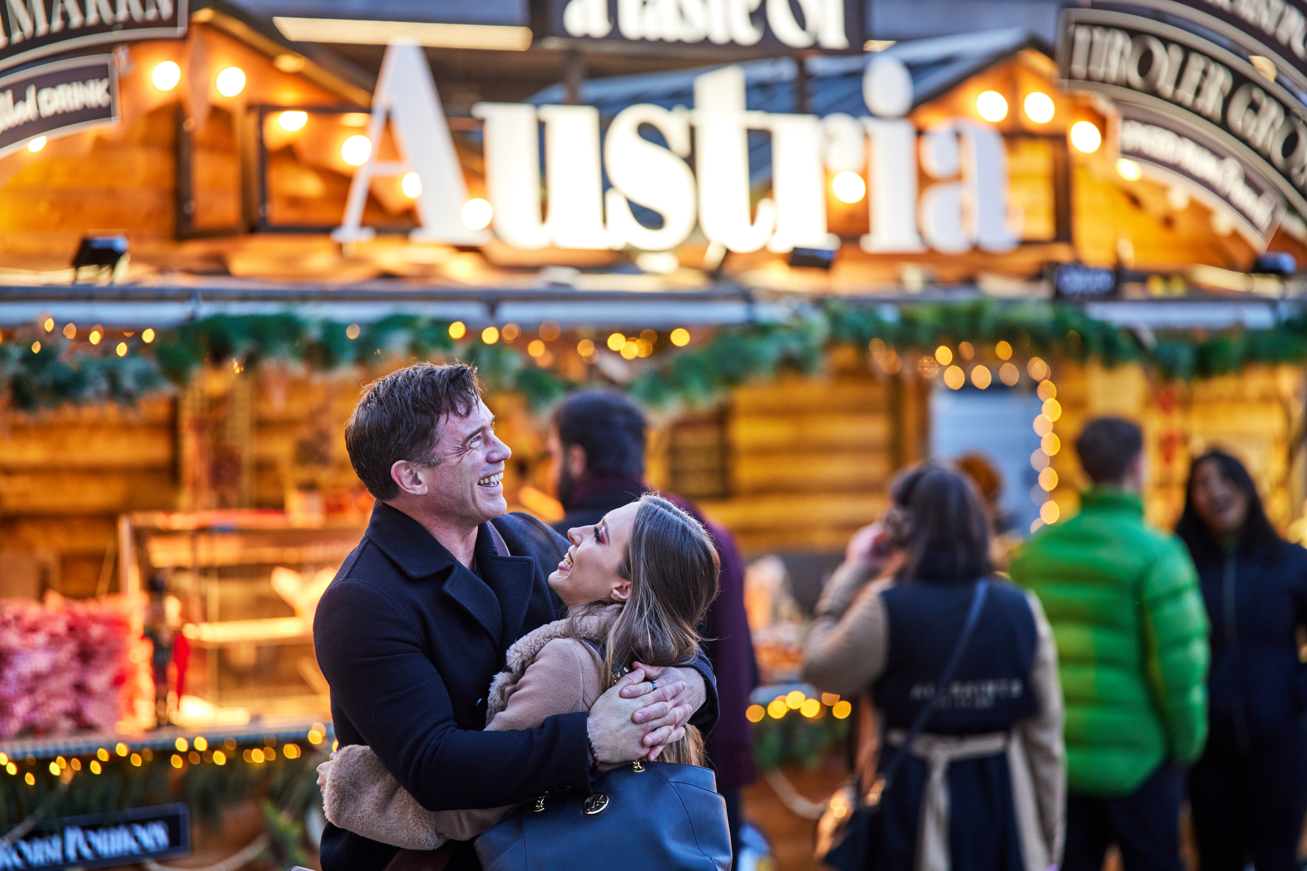 Outdoors with the bright twinkling lights of a market stall behind them, a man and a woman smiling at each other as they hug.