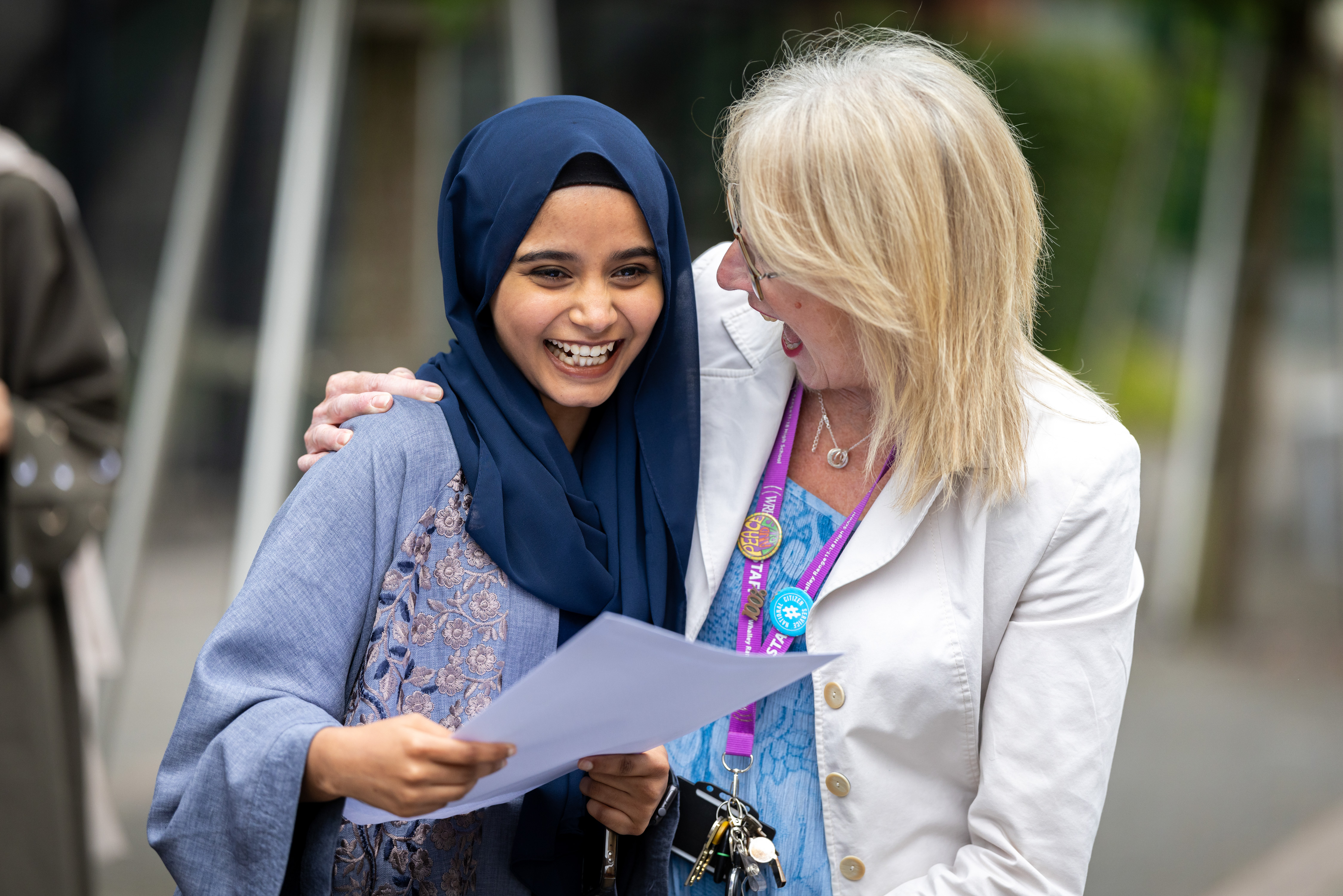 Teenage girl smiling holding piece of paper in her hand, as a woman looks smiling at her with her arm around her Teenage girl smiling holding piece of paper in her hand, as a woman looks smiling at her with her arm around her