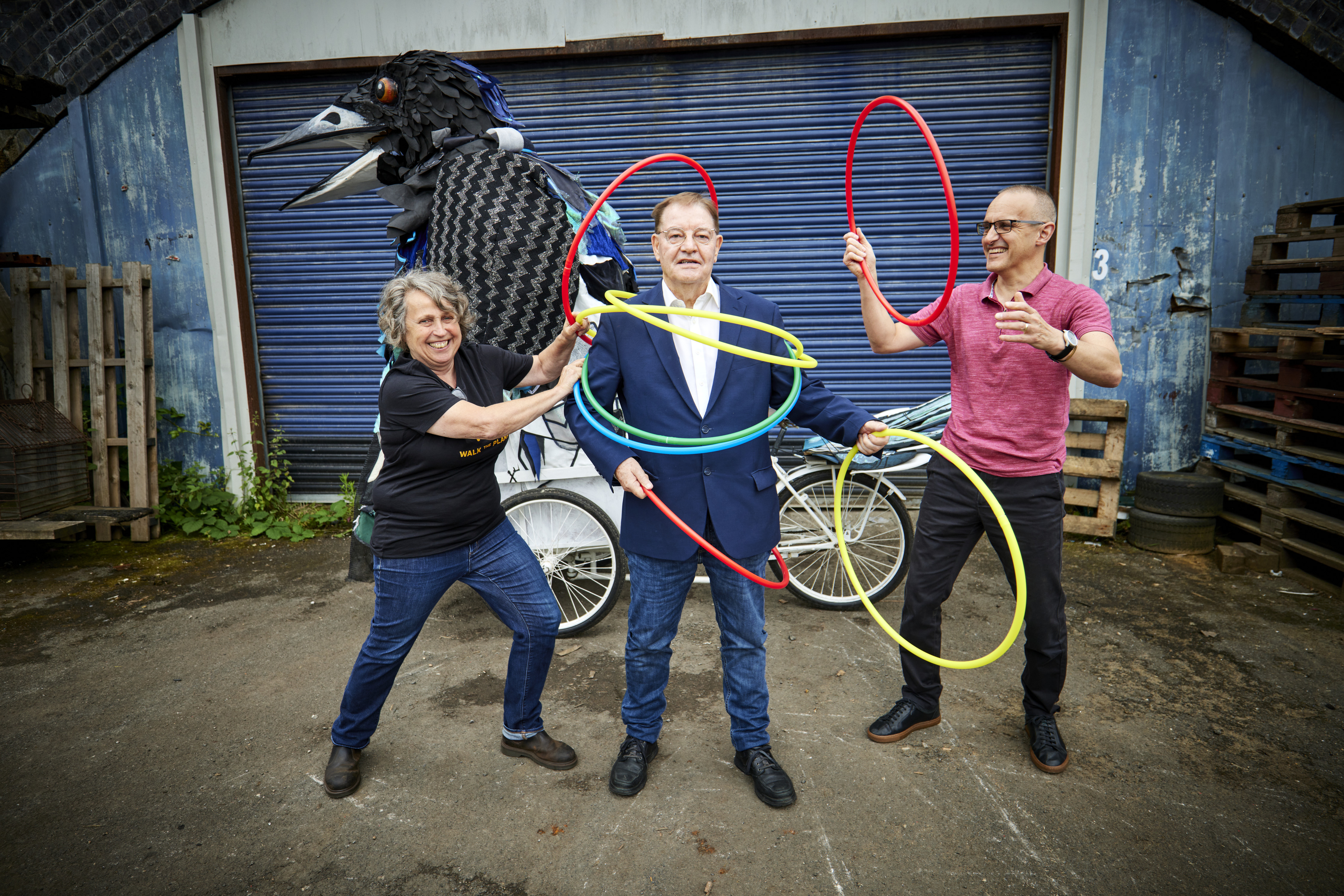 Three laughing adults in a row, the two adults on either end are putting big hula hoops over the head of the person in the middle.