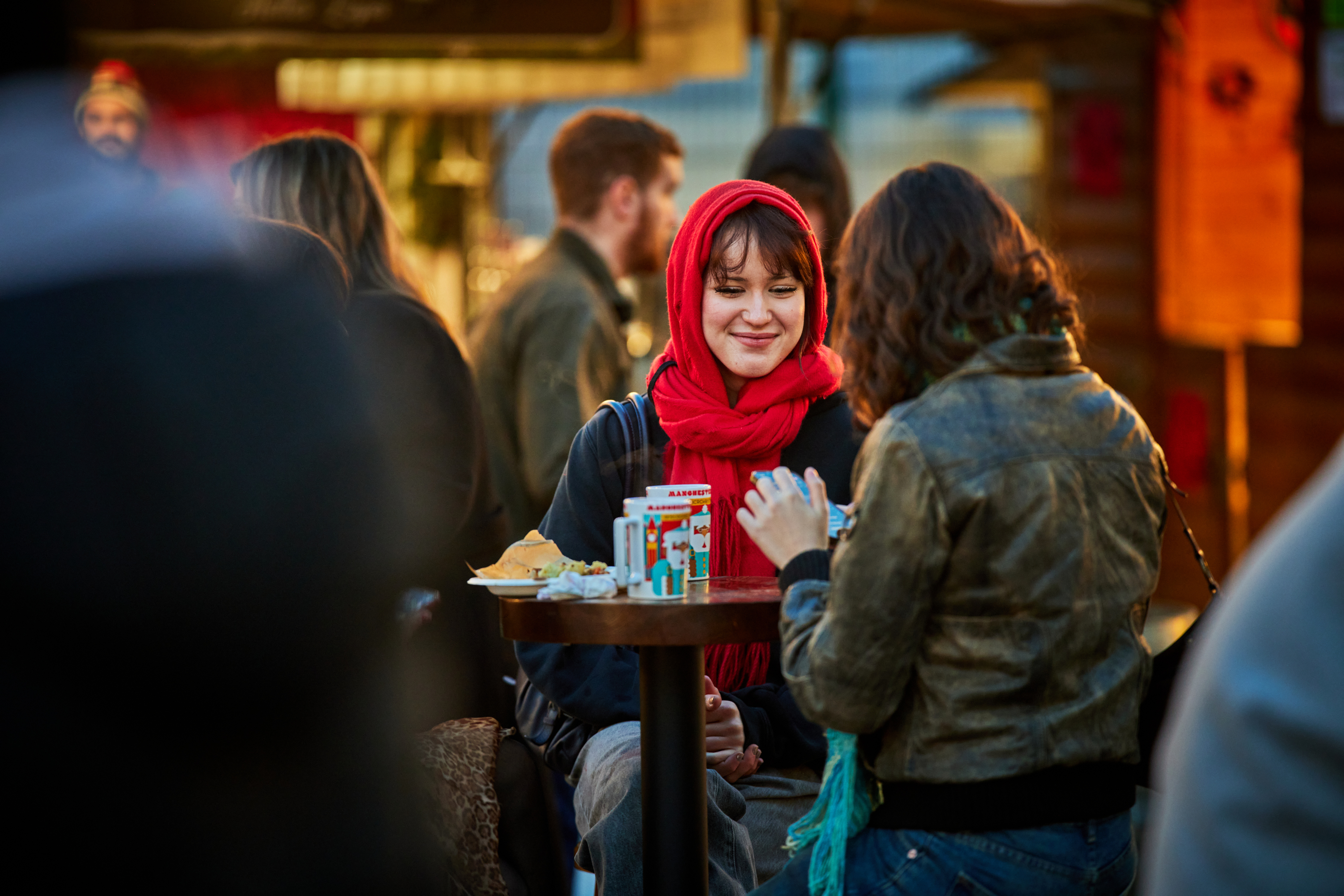 A young smiling woman wearing a red headscarf sits at a table outside with another person