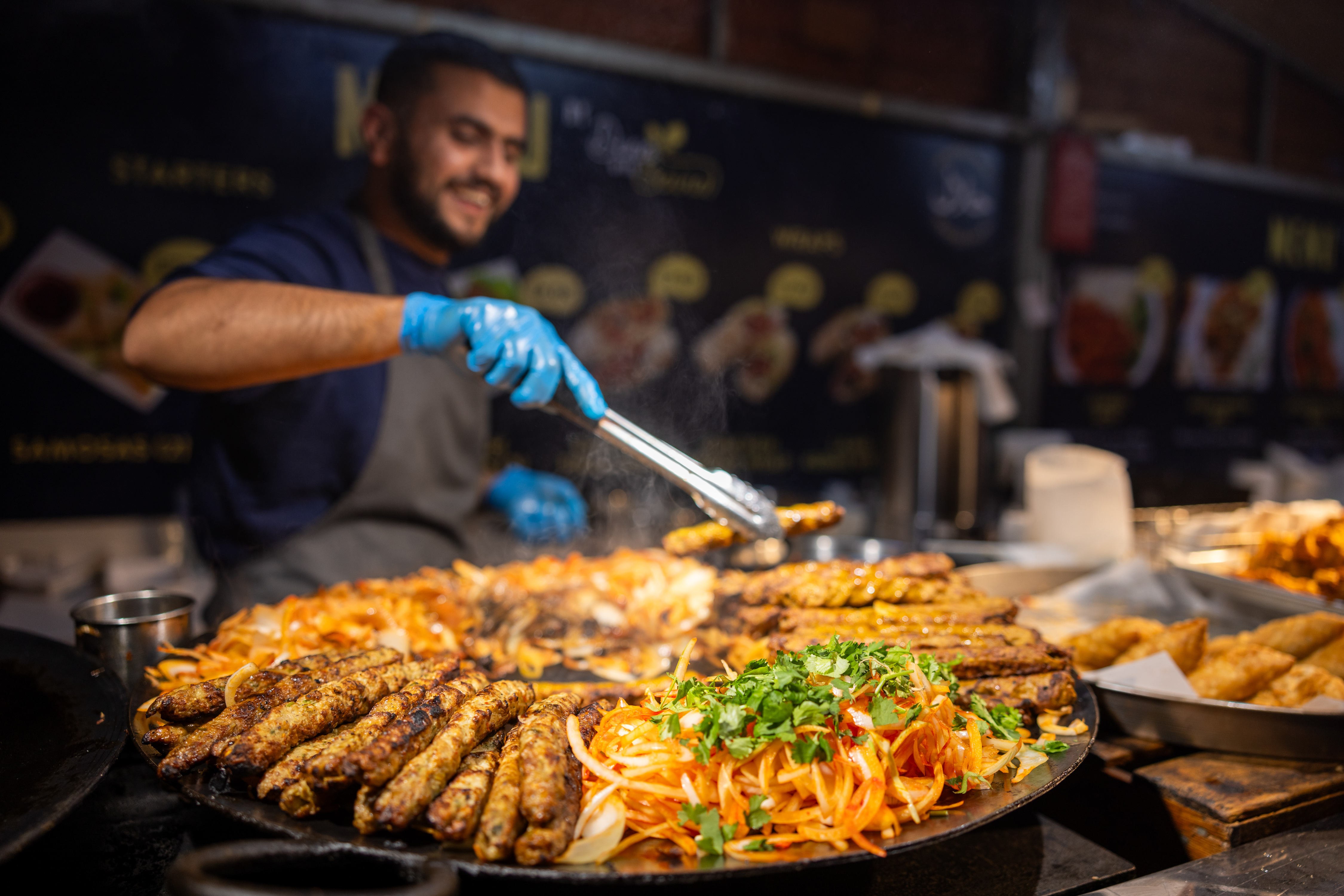 An enormous pan of food being cooked outside in a market stall with the chef standing behind stirring the food 